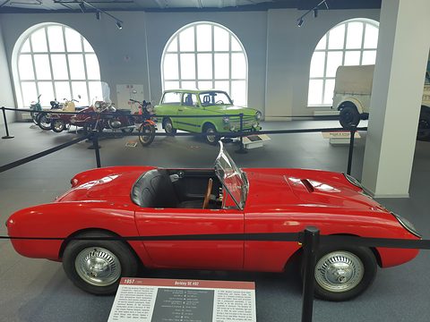Red open two-seater in profile in a bright hall with arched windows, a caption board before it. Opens a larger view