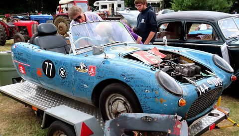 Scuffed blue three-wheeler covered in signatures and race numbers on a car trailer, men behind. Opens a larger view