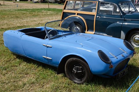 Blue open three-wheeler on grass beside a wood-framed estate car. Opens a larger view