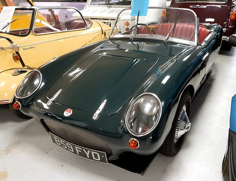 Dark green open three-wheeler on a pale floor, seen close from the front right. Opens a larger view