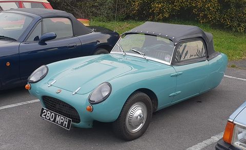 Turquoise three-wheeler with a black hood parked in a car park, seen from the front left. Opens a larger view