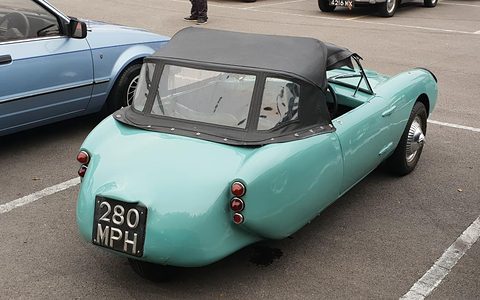 Turquoise three-wheeler from behind in a car park, a black hood over the cockpit. Opens a larger view