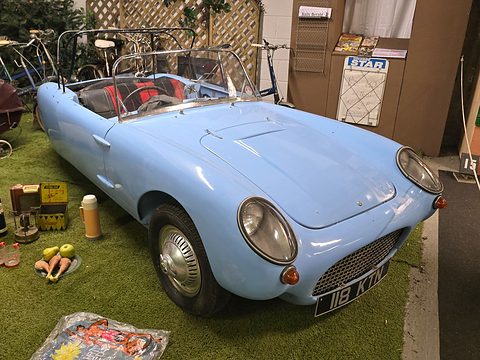 Pale blue open three-wheeler on artificial grass among picnic props, seen from the front right. Opens a larger view