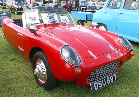 Red open three-wheeler with a display card on its screen, on grass by a pale blue car. Opens a larger view