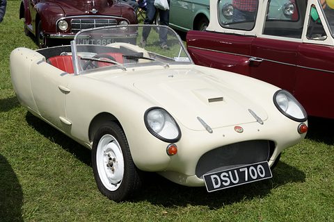 Cream open three-wheeler on grass, seen from the front right, red cars behind. Opens a larger view