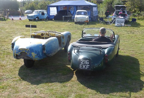 Bare, battered blue three-wheeler body next to a complete green one on grass, tents behind. Opens a larger view