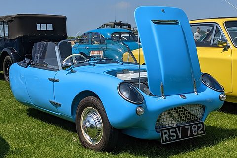Bright blue three-wheeler with its whole nose tilted up on grass, other cars behind. Opens a larger view