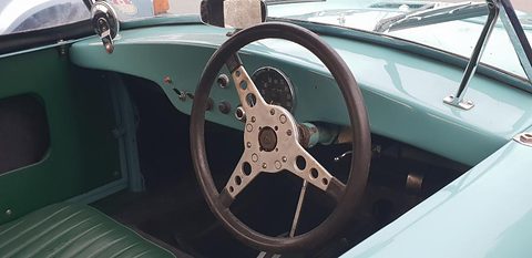 Wood-rimmed steering wheel with drilled spokes in front of a turquoise dashboard and round dials. Opens a larger view