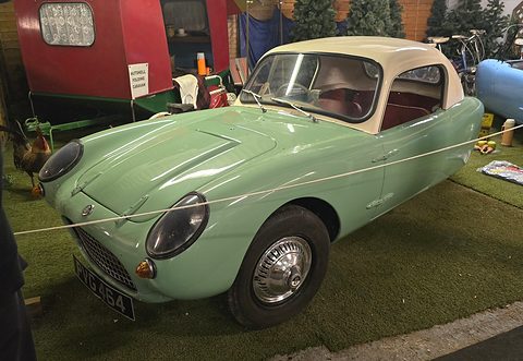 Pale green three-wheeler with a cream hardtop on artificial grass behind a rope. Opens a larger view