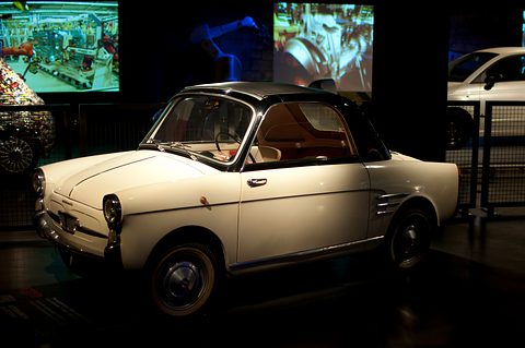 Cream small coupé with a black roof and chrome side vents, lit in a dark gallery with factory films on screens behind. Opens a larger view