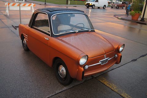 Burnt-orange small coupé with a black roof and chrome trim, parked on a wet street with barriers behind. Opens a larger view