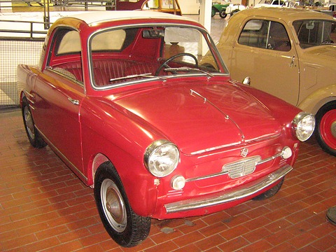 Red small coupé with a cream roof panel and red seats, on a brick museum floor beside a tan Fiat Topolino. Opens a larger view
