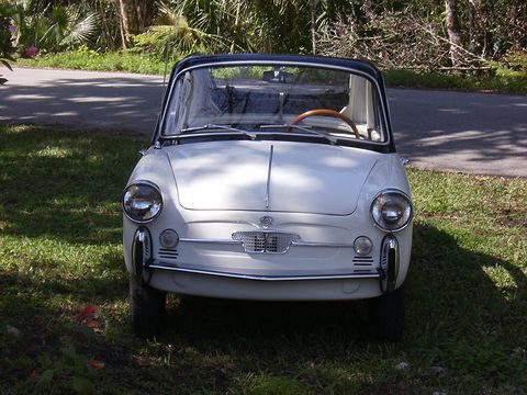 Front of a white small coupé with a black roof, round headlamps and a small chrome grille, parked on grass. Opens a larger view