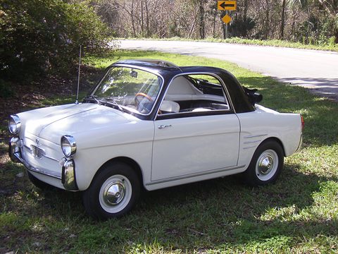 White small coupé with a black canvas roof and chrome bumpers, parked on grass by a curving road. Opens a larger view