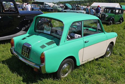 Mint-green and cream small saloon seen from the rear left on grass, a black vintage car and tents behind. Opens a larger view