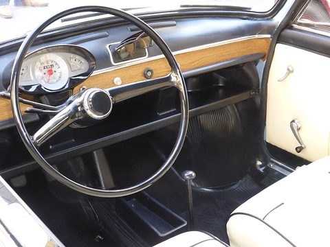 Black dashboard with a wood-grain strip, a round speedometer and a large thin chrome-spoked steering wheel.