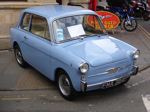 Pale blue small saloon with a chrome grille and British plate, parked on a pavement before a motorcycle club sign. Opens a larger view