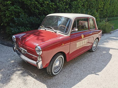 Dark red small saloon with a cream roof and gold lettering on its door, parked on gravel before a hedge. Opens a larger view