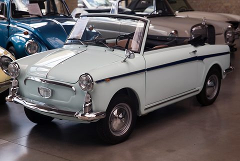Pale grey-white open two-seater with a dark blue waist stripe and a sale card on its windscreen, in a showroom. Opens a larger view