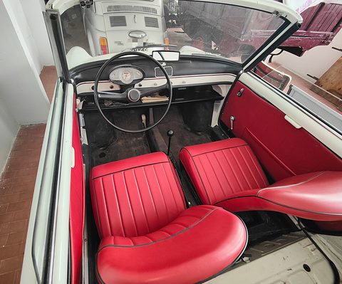 Looking down into a white open car with two red pleated seats, a thin steering wheel and a round speedometer. Opens a larger view