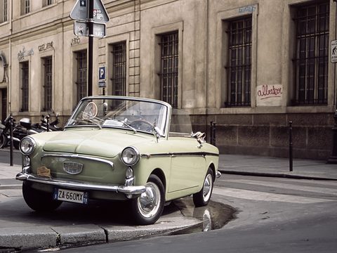 Pale green open two-seater with chrome bumpers and Italian plates, parked on a kerb before a stone building.