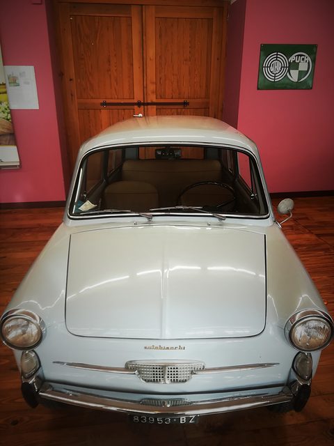 Front of a white small saloon with an Autobianchi script and small grille, against a red wall and wooden door. Opens a larger view
