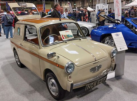 Cream small van with a wood-coloured stripe, its rear door raised, on a show stand with visitors around. Opens a larger view