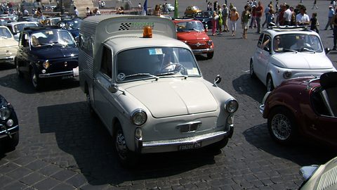 Pale grey small van with a tall box body and an orange beacon, surrounded by Fiat 500s and people on cobbles. Opens a larger view