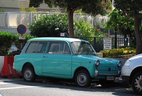 Turquoise small estate car with a long glass cabin, parked on a street beside red and white barriers and trees. Opens a larger view
