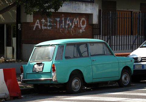 Turquoise small estate car seen from the rear left, graffiti on the wall behind and a white car in front.