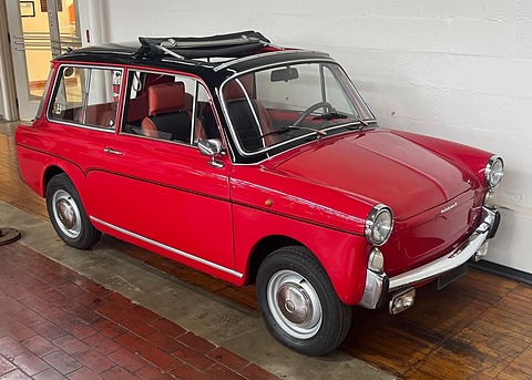 Red small estate with a black roof whose long canvas sunroof is rolled back, against a white brick wall. Opens a larger view