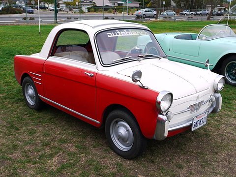 Small coupé with a red body, white roof and bonnet, and an autobianchi sun strip, parked on grass. Opens a larger view