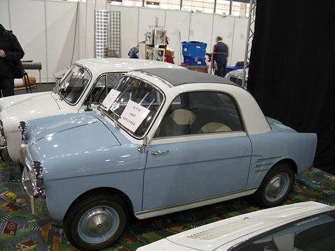 Pale blue small coupé with a white roof and chrome side vents on patterned carpet, another Bianchina behind. Opens a larger view
