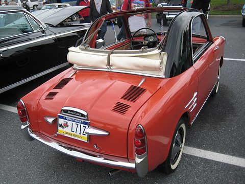 Coral-red small coupé from the rear, its canvas roof furled behind the seats, a Pennsylvania antique plate on the tail.