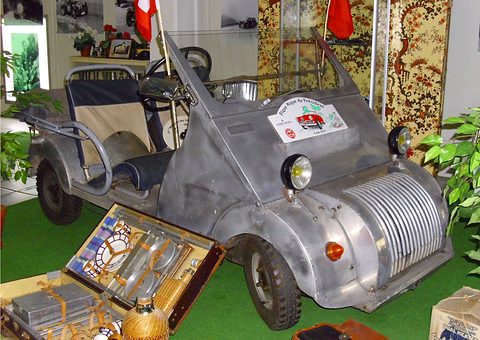 Bare aluminium open microcar with a ribbed nose and rally plaque, a picnic case open beside it.