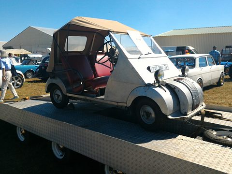 Aluminium microcar with a beige canvas hood and red interior on a flatbed trailer under blue sky.