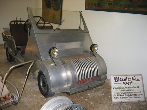 Unpainted aluminium open microcar with a ribbed grille and a hand-lettered museum sign in front. Opens a larger view