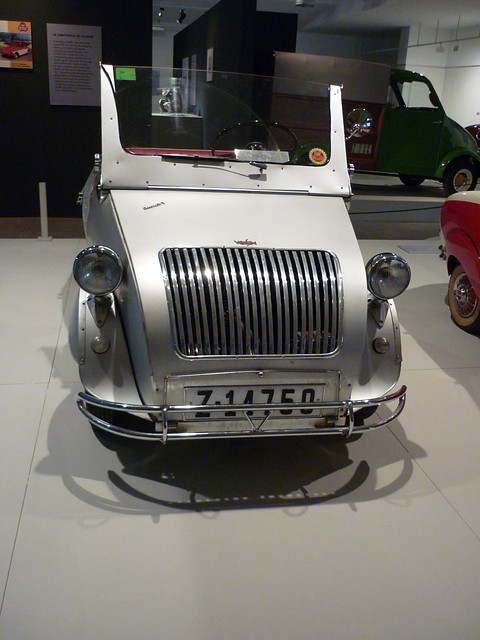 Front of a silver open car with a tall curved grille of vertical bars, two pod headlamps and a raised windscreen, in a museum. Opens a larger view