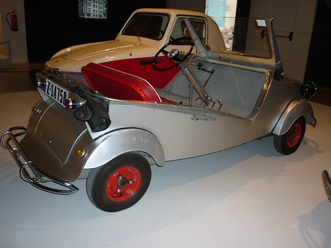 Silver open two-seater from the left, low cut-away sides, red seat and red wheels, a cream microcar behind. Opens a larger view