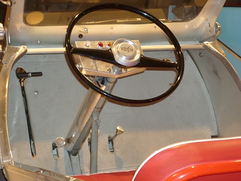 Large black thin-rimmed steering wheel above a plain aluminium floor and scuttle, a gear lever and two pedals. Opens a larger view
