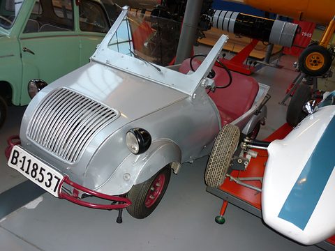 Silver doorless two-seater with a curved vertical-bar grille and red bumper, windscreen raised, among museum exhibits. Opens a larger view