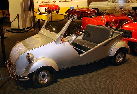 Silver doorless car with a grey bench seat and orange indicator, parked on a dark carpet among red sports cars. Opens a larger view