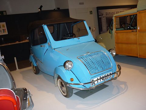 Light blue small car with a curved vertical-bar grille, pod headlamps and a black hood, beside a wooden-bodied van. Opens a larger view