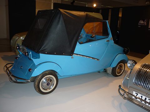 Light blue small open car with a black canvas hood, tubular rear bumper and whitewall tyres, seen from the rear left. Opens a larger view