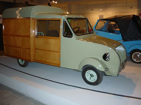 Side view of a pale green van cab joined to a tall wooden box body, a blue hooded microcar behind. Opens a larger view