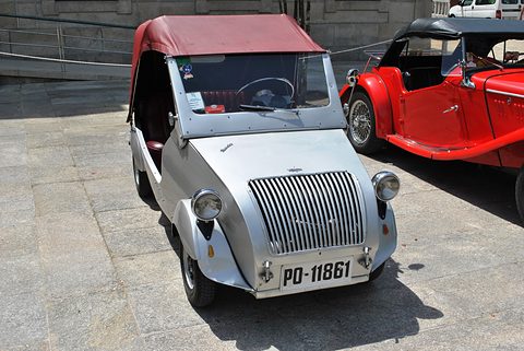 Silver small car with a red canvas hood and vertical-bar grille on a stone-paved square, a red roadster behind. Opens a larger view