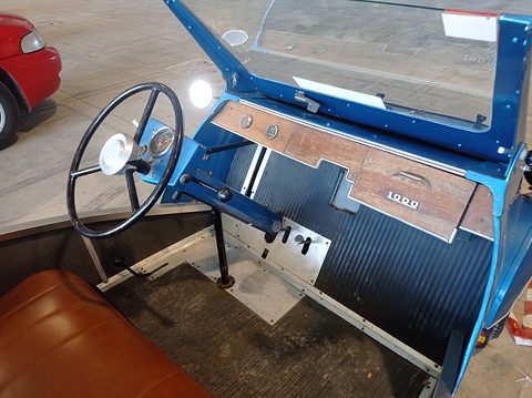 Blue painted scuttle with a wooden facia strip, thin steering wheel, gear lever on the floor and a tan bench seat. Opens a larger view