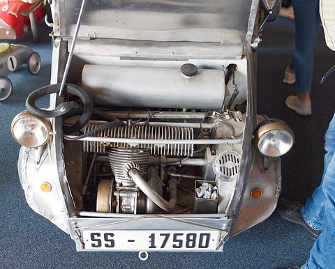 Open nose of a silver car showing a finned single-cylinder engine and its silencer between two headlamps, plate SS-17580. Opens a larger view