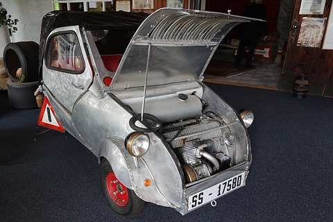 Weathered silver small car from the front left with the whole nose raised, exposing a finned engine, a warning triangle beside it. Opens a larger view