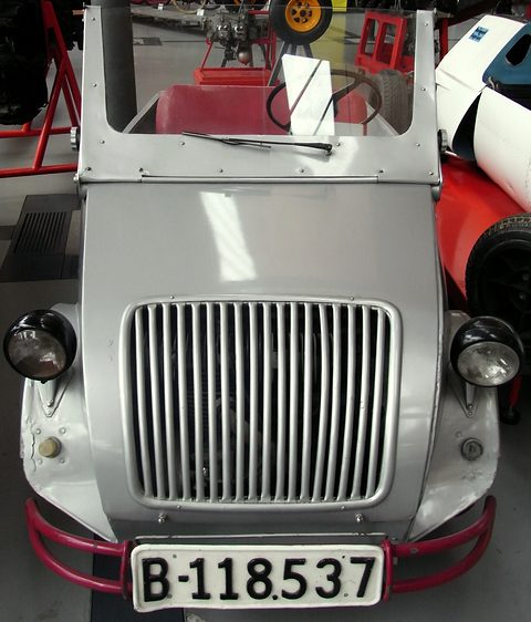 Head-on view of a silver car with a tall grille of vertical bars, pod headlamps, a red bumper and plate B-118537. Opens a larger view